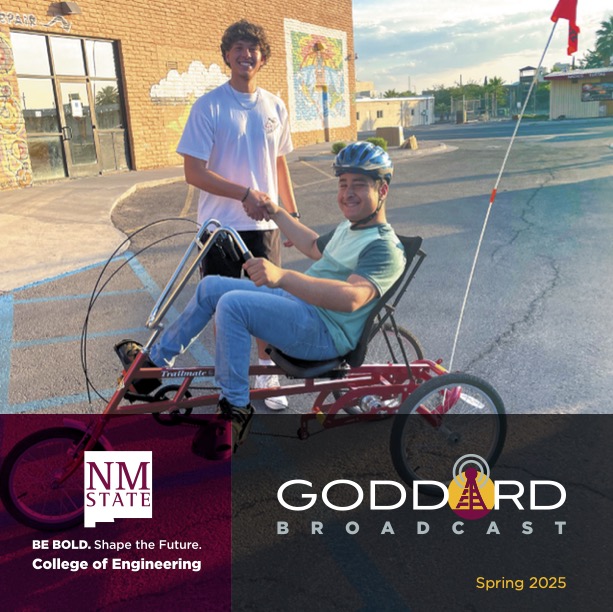 Student wearing a helmet sits in a recumbent tricycle and shakes hands with another student in a parking lot outside a brick building.